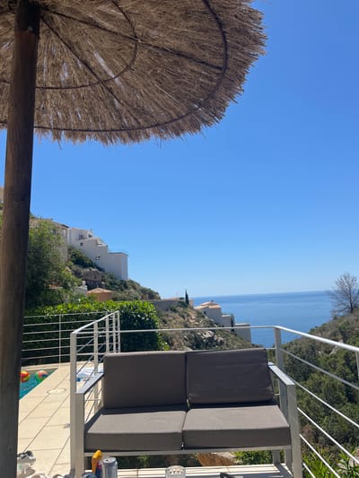 View from a terrace overlooking the Mediterranean Sea in El Poble Nou de Benitatxell, Spain. A thatched parasol is on the left. A gray outdoor sofa sits behind a white metal railing. Below is a swimming pool with inflatable balls. White buildings are on a hill, against a clear blue sky.