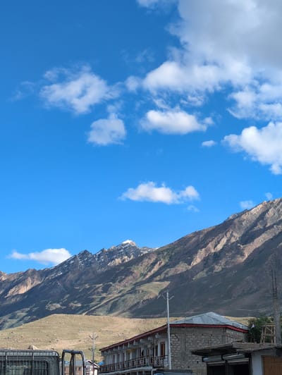The image depicts a mountainous landscape under a bright blue sky dotted with white clouds. In the foreground, there is a building with a red-trimmed roof and stone walls. A metal fence and parts of a vehicle are visible on the left. A utility pole stands near the building. The middle ground shows a barren, sloping hillside leading up to a majestic mountain range, whose peaks are dusted with snow. The mountain slopes are characterized by dry vegetation and shadows. The scene is likely a street view in or near the city of Padum, India, given the architecture and mountainous terrain.