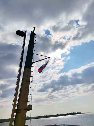 A low-angle shot captures a tall, industrial-looking mast against a cloudy sky. The mast is dark in silhouette, with a round object, possibly a light, at its top. A Russian flag, displaying white, blue, and red horizontal stripes, is attached to the mast by a thin rope, fluttering in the breeze. The mast appears to be part of a ship or a structure on the water. The background shows a body of water, likely a river or lake, with a distant shoreline featuring trees. The sun peeks through the clouds, creating bright spots and casting a shimmering effect on the water. The scene evokes a sense of maritime activity and a vast, open sky. The location is Krokhino, Russia.