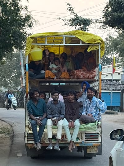 A light commercial vehicle, identified by the license plate "KA39B 2775," transports a substantial group of individuals on a road in Chikkarampur, India. The vehicle's cargo bed is fitted with a metal frame and covered by a yellow tarp.

Approximately eight to ten individuals, primarily women and children, are seated within the upper, tarp-covered section of the cargo bed. Several of these individuals are smiling and waving. One person balances a yellow plastic container on their head.

Positioned on the lower section, at the rear edge of the vehicle, are at least six adult males and one young girl. One male on the far left is holding a smartphone. Multiple individuals in this lower group are also smiling.

The surrounding environment includes a paved road with a dirt shoulder and green trees on the left, with power lines visible overhead. A white scooter carrying two male riders is observed in the middle distance on the road. To the right, a blue structure and a flagpole displaying a red and yellow flag are visible. A portion of a white vehicle, including its side mirror, occupies the extreme right foreground.