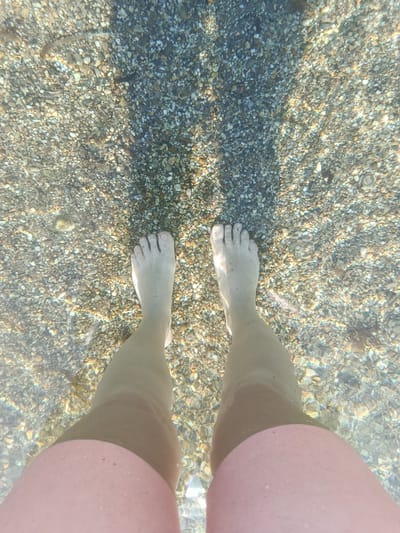 The image captures an overhead, underwater perspective of a person's legs and feet submerged in clear, shallow water, likely at a beach in Águilas, Spain. The scene shows the individual standing on a seabed densely covered with small, varied-colored pebbles and rocks.
The water is remarkably transparent, allowing for distinct visibility of the individual stones and sand on the bottom. Sunlight penetrates the water, creating vibrant, shimmering patterns and refractions across the seabed, indicating a sunny day. The person's bare feet are positioned firmly on the pebbly surface, with their lower legs extending upwards. Pink swim shorts are visible at the very bottom of the frame, indicating the person's attire and that the water level reaches approximately their upper thighs or knees. Shadows, possibly cast by the person's body or surrounding elements, are faintly visible on the seabed. The overall impression conveys a peaceful and clear aquatic environment. There is no visible text in the image.