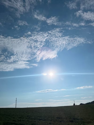 A wide-angle, low-angle shot captures a sunlit sky above a dark, grassy field. The sun, a bright, starburst-like orb, is positioned slightly left of the center of the frame, emitting a strong lens flare that creates a horizontal streak of light across the image. Wispy, white clouds are scattered across the brilliant blue sky, with some appearing denser and more textured than others. In the middle ground, a dark, solitary utility pole stands to the left of the frame, supporting power lines that stretch horizontally towards the right. Another smaller utility pole is visible in the distance on the right side of the frame, and a line of trees forms a dark silhouette against the horizon on the far right. The foreground is dominated by a dark, out-of-focus expanse of grass, suggesting a field under a bright sky. The scene appears to be set in a rural, open landscape, possibly on the outskirts of Vertelka, Ukraine, characterized by its agricultural fields and utility infrastructure against a dramatic sky.