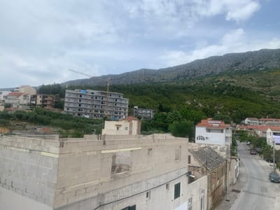 The image shows a view from a rooftop looking down a paved street in Dugi Rat, Croatia. In the foreground, a partially constructed building is visible, its concrete blocks laid bare. The street descends towards the bottom of the frame, lined with buildings on both sides. On the left, a tall, multi-story building under construction dominates the mid-ground, with a crane visible above it. The building is seemingly unfinished, with no windows or doors visible. The background is dominated by a verdant hillside, with trees and brush covering the slopes. A large, rocky mountain ridge can be seen in the distance, forming the upper skyline. The sky is partly cloudy, with patches of blue sky visible. There are no people visible in the scene.