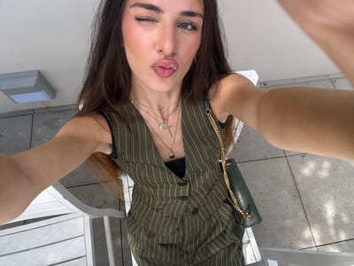 In Hallandale Beach, Florida, a young woman is captured in a selfie outdoors, seated on a white chair. She is wearing a green pinstripe vest over a black top and accessorized with multiple gold necklaces, one featuring a cross pendant. A green crossbody bag with a gold chain strap is visible on her right side. The woman is winking and puckering her lips, looking towards the camera. The background shows a light-colored ceiling, a grey outdoor light fixture, and grey paving stones.