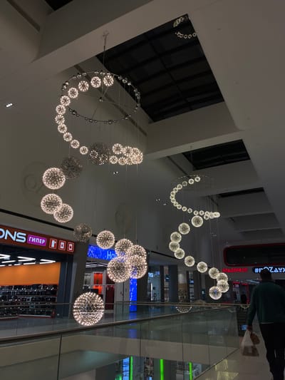 The image displays the interior of a large, modern building, likely a shopping mall, characterized by its high ceilings and elaborate decorative lighting. Numerous spherical chandeliers, crafted from many small, glittering lights, hang suspended, forming dynamic patterns that include elongated waves and complete circles. These fixtures provide the primary illumination for the space.
One person is visible in the upper right quadrant of the image, seen from behind. They are dressed in a dark green top and dark pants, and are carrying a light-colored bag, suggesting they are a shopper or visitor moving through the facility. The perspective implies the person is on an upper level, looking down or across the expansive interior.
Glass railings are prominent features, indicating a multi-story structure with various walkways or balconies overlooking the main area. In the background, multiple shop fronts and displays are discernible, reinforcing the commercial nature of the setting. The lighting is entirely artificial, creating a bright yet focused ambiance typical of an indoor public space, irrespective of the time of day outside.
Several Russian brand names are visible on store signage. These include "М.ВИДЕО" (M.VIDEO) and "#Эльдорадо" (Eldorado), both identifiable as major electronics retailers. Another sign clearly shows the word "гипер" (giper), which is likely an abbreviation for "гипермаркет" (hypermarket). The scene captures a typical moment within a modern commercial hub in Tliustenkhabl', Russia.