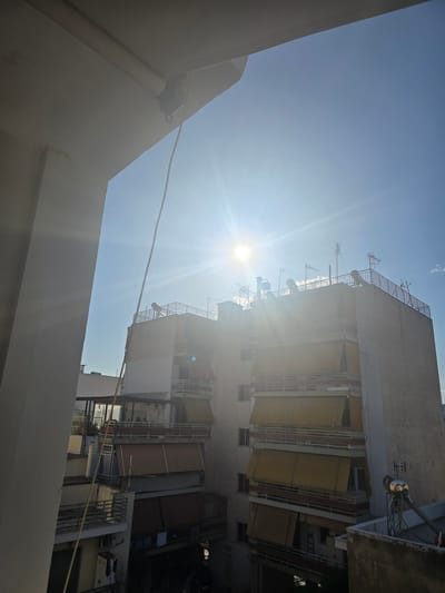 The image shows a view of apartment buildings in Agii Anargiri, Greece, on a sunny day. The sun is bright and positioned high in the sky, creating lens flare and strong highlights. A prominent white building facade, possibly part of a balcony or overhang, is in the foreground on the left, partially obscuring the view of the sky and the buildings beyond. A thin wire hangs vertically from the overhang into the frame.
The main subject is a multi-story apartment building with multiple balconies. Many of the balconies are equipped with retractable awnings, most of which are extended and are a yellowish-tan color. Some awnings are retracted or not visible. The building has white railings on its balconies and appears to be constructed of concrete. The rooftop of the building is visible and is equipped with several antennas, including a large satellite dish and smaller aerials. There are also what appear to be solar water heaters on the rooftop. Another, slightly shorter building is visible behind and to the left of the main apartment building. The sky is clear and blue, with a few wispy clouds visible. The overall scene depicts an urban residential environment. No people are visible in the image.