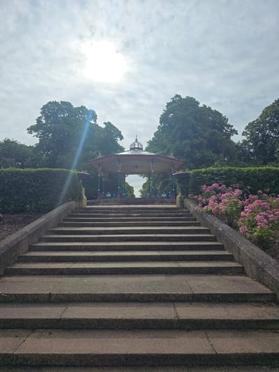 This image captures a picturesque outdoor scene, oriented vertically. In the foreground, a prominent stone staircase with weathered steps ascends, bordered by a concrete railing. To the left of the stairs, a meticulously manicured hedge forms a verdant boundary. Beyond the hedge, a charming bandstand with a red roof and blue accents is visible, nestled amongst lush green trees. Pink flowers add a splash of color to the scene, blooming near the top of the hedge and railing. The sky above is cloudy, with the sun shining brightly through, creating a lens flare effect. The overall impression is one of a peaceful park setting, likely on a pleasant day, possibly in the late morning or early afternoon, given the bright sunshine. The location appears to be a public garden or park in Stockton-on-Tees, United Kingdom, given the contextual information. There is no visible text in the image. The scene evokes a sense of tranquility and natural beauty.