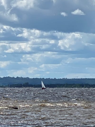 This is a photograph of a sailboat on a body of water with a cloudy sky and a tree-lined distant shore. The water is choppy, with sunlight glinting off the waves. The sailboat has a white sail and a dark hull, and it is positioned in the middle of the frame. The sky is filled with dark, dramatic clouds, suggesting an overcast day. The distant shore is a dark green line of trees, and there appears to be a faint structure on the horizon. The scene appears to be a natural landscape, possibly a lake or a river, with no immediate signs of urban development. The photograph is taken from a low vantage point, looking out over the water. No people or other boats are visible. Based on the provided crop hints, this scene could be located in the city of Leninsky, Russia, likely on a waterfront area.