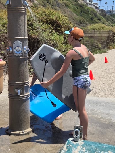 The image depicts a young person, likely a girl, standing at a public shower station on a beach in San Clemente, California. She is holding a grey boogie board with a black string attached and a blue boogie board. Water is running from the shower head onto the boards, suggesting she is rinsing them. She is wearing a green tank top and patterned shorts, and a baseball cap. The shower station is a large, cylindrical structure with chrome fixtures. Behind her, a sandy beach slopes upwards to a grassy hillside with buildings visible in the distance. Orange traffic cones are scattered on the beach. The ground around the shower is wet and sandy. The overall scene is typical of a sunny day at the beach.