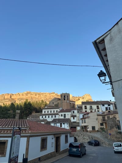 This is a horizontal, eye-level, full shot of the town of Alpuente, Spain, as seen from a street corner. The sky is a clear, bright blue. In the foreground, a white building with a stone foundation and a terracotta tiled roof runs along the bottom right of the image. A street lamp hangs from the upper right corner of the building.

To the left, a street slopes down to the right, with several parked cars visible. A dark blue minivan is parked in the lower left of the street, with license plate 2639 HLR. A black sedan is parked further up the street, and a white van is parked on the right.

The middle ground of the image features a cluster of multi-story buildings with terracotta tiled roofs. Some buildings are white, while others are painted in pastel colors like cream and light yellow. One building has a tall bell tower with a clock face.

In the background, a large, rocky cliff face rises behind the town. The cliff is bathed in golden sunlight, contrasting with the shadows cast on the town below. A line of green trees runs along the base of the cliff. A thin black wire stretches across the upper portion of the image, likely a power line or telephone wire. A signpost to the left of the foreground building has text in Spanish and an image of the town.
