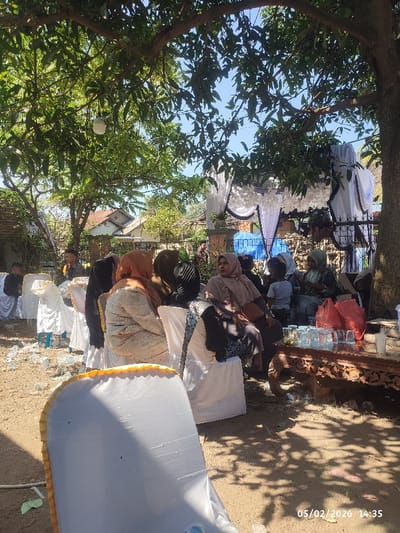 The image captures an outdoor social gathering under the canopy of large, leafy trees, suggesting a sunny day with natural shade. The scene appears to be set in a residential or community area, possibly a backyard, with houses visible in the background through the foliage.

Numerous people, mostly adults and predominantly women, are seated on white chairs with fabric covers. Some of the chair covers have a yellow or gold trim along the top edge. Many of the women are wearing headscarves (hijabs). A child is also visible among the adults. The individuals appear relaxed, some engaged in conversation while others are looking forward or to the side.

To the right side of the frame, a low, intricately carved dark wooden table holds several clear drinking glasses stacked together and two red plastic bags. Further back, a decorative structure in white and black is visible, resembling an arch or a backdrop, possibly for a special event like a wedding or a celebration, adorned with white ruffles and dark accents.

The ground beneath the chairs is bare earth, and there is some scattered debris, including plastic bottles and wrappers. The strong sunlight filtering through the trees creates distinct shadows on the ground and dappled light on some of the chairs and people.

A timestamp "05/02/2026 14:35" is digitally embedded in the bottom right corner of the image.