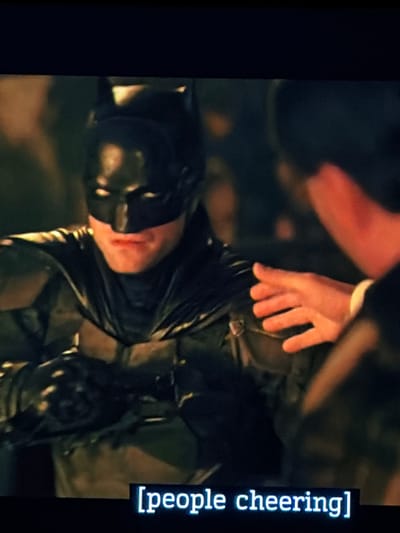 The image shows Batman in his suit, lying down with a hand reaching towards him. The scene appears dark and possibly injured. Subtitles on the right side read "[people cheering]" suggesting an off-screen reaction to the scene. The quality is grainy, indicating it may be a still from a video.