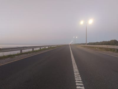 The image depicts a straight, paved road with a yellow line on the right shoulder and a white dashed line in the center, receding into the distance. The road is flanked by a metal guardrail on the left, separating it from a body of water. On the right side of the road, a line of trees is visible beyond the guardrail. Several streetlights are spaced along the road, some of which are illuminated, casting a bright glow. In the far distance, a car's headlights are visible. The sky is overcast and a pale grey, suggesting either dawn or dusk. The overall atmosphere is serene and slightly hazy. The location is identified as Zavolzhye, Russia.