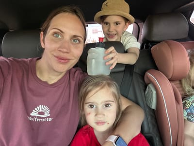 The image depicts a selfie taken inside a vehicle. A woman and two children are visible. The woman is wearing a maroon t-shirt with white text on the chest, positioned on the right side of the frame. A young girl wearing a red shirt and a smart watch is centered at the top, and a younger boy in a straw hat and white shirt is at the bottom. The boy is holding a translucent plastic water bottle. The background shows dark leather car seats and a glimpse of the outside through the car window. The location is inferred to be the city of Ula, Turkey, based on the assumption that the photograph was taken there. The image is rotated 180 degrees, with the bottom of the car facing up.