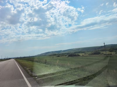 The image depicts a rural roadside scene on a sunny day. A paved road with a white line marking its edge curves to the left, bordered by a grassy shoulder. Beyond the shoulder, a vast, green field stretches towards a series of rolling hills in the distance. Several utility poles with wires are visible along the roadside and across the field. In the far distance, a line of trees marks the horizon. The sky is predominantly blue with scattered white cumulus clouds. The scene appears to be in the vicinity of the village of Pokrovka, Russia, characterized by its open fields and agricultural landscape. No people or specific structures of note are identifiable within the frame.