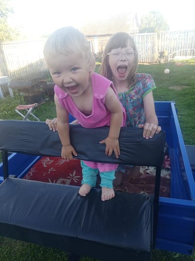 In Buraly, Russia, two young girls are depicted in the back of a blue utility trailer. The younger girl, with blonde hair, is leaning forward, smiling broadly, and wearing a pink sleeveless shirt and turquoise pants. Her hands are on the black padded seat of the trailer. The older girl, with brown hair and glasses, is behind her, also with her mouth open in a shout or laugh. She is wearing a colorful patterned dress and her hands are resting on the same seat. The trailer contains blankets, one red with a white pattern and another brown. The background shows a wooden fence, a building, and green grass, suggesting a rural or suburban setting.