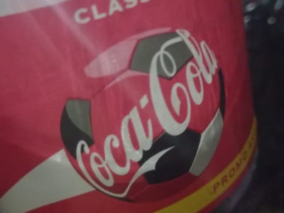 This media file features a close-up view of a red Coca-Cola can with a soccer ball graphic. The can is partially curved, suggesting it is a cylindrical object. The dominant color is red, with the iconic white Coca-Cola logo prominently displayed across the soccer ball design. The soccer ball itself is depicted with black and white hexagonal panels, a common representation. Above the soccer ball and logo, the word "CLASS" is partially visible in white text. Along the bottom edge of the can, a yellow and red striped band contains yellow text that reads "PROMO". The background is dark and out of focus, providing little context about the surrounding environment. The lighting is dim, creating some glare and reflections on the can's surface. There are no people or animals visible, and the scene appears to be a still image of a product. The context of "Yorkatay, Uzbekistan" is provided, but there are no visual cues within the image itself to confirm this location. The image focuses on the product's branding and design.