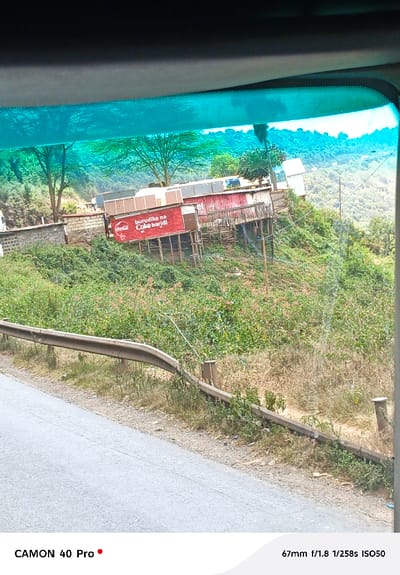A roadside scene with a commercial structure advertising Coca-Cola in what appears to be Swahili, "burudika na Coke baridi," translating to "refresh with cold Coke." The building is perched on a hillside amidst lush vegetation, with a view of rolling hills in the background. A damaged guardrail runs alongside the road, and the photograph appears to be taken from inside a vehicle, as evidenced by the tinted windshield. The image is captured with a CAMON 40 Pro, using a 67mm lens at f/1.8, with a shutter speed of 1/258s, and an ISO of 50.