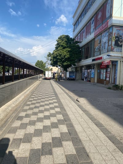 The image depicts a street scene in Parkent, Uzbekistan, on a sunny day. The foreground is dominated by a checkered tiled sidewalk with alternating dark gray and light gray rectangular tiles. To the left, a long, covered walkway with a metal roof supported by wooden beams runs parallel to the sidewalk, with a dark gray stone wall and a drainage channel along its base. Beyond the walkway, trees and a white van are visible.
To the right of the sidewalk is a multi-story building with a white facade, featuring numerous windows and signage. The signage includes advertisements for "ABROR AVTO TEST" and other services, with phone numbers displayed. Several shops are visible at the ground level of the building, including one with a red awning that reads "ABROR AVTO TEST mobiliz". A few people are walking on the sidewalk in front of the building, and a white truck is parked further down the street. The sky is blue with scattered white clouds.
