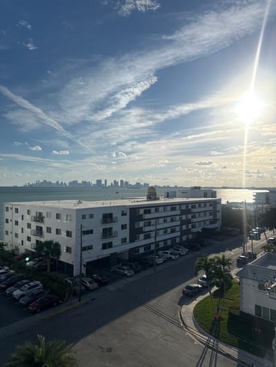 The image displays a high-angle view of a coastal cityscape on a sunny day. A tall, white apartment building dominates the foreground, lined with parked cars. Beyond the building is a body of water, separating it from a distant skyline. The sky is blue with scattered clouds, with the sun creating a bright glare. A street runs along the building, complete with a sidewalk and some greenery. The view is from an elevated position, possibly a higher floor of another building.