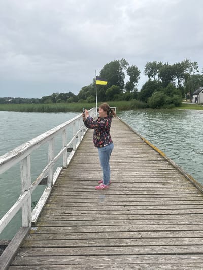 A person stands on a wooden pier, taking a photograph with their mobile phone. The pier extends into a body of water, with reeds lining the distant shore. Trees and buildings are visible on the far side of the water, under a cloudy sky. A yellow and black flag flies on a flagpole in the distance. The person is wearing a colorful patterned jacket, blue jeans, and pink sneakers. The pier is weathered, with visible grain and wear on the wooden planks and white painted railings. The water is choppy, indicating wind. The scene appears to be a serene natural setting.