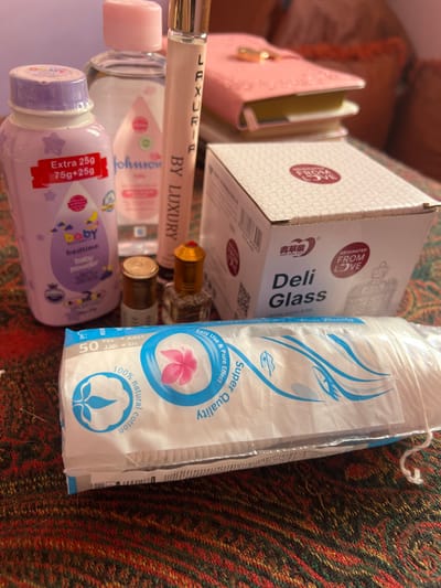 This close-up image, taken in Ksar-el-Kebir, Morocco, depicts a collection of personal care products and accessories arranged on a patterned textile surface. The background is blurred, showing soft furnishings in muted tones.

Prominently displayed are:
*   A lavender-capped bottle of "baby bedtime baby powder" labeled "EXTRA 25g 75g+25g," situated on the left.
*   Behind it, a clear bottle of "johnson's baby" oil with a pink cap.
*   A tall, slender silver-capped perfume rollerball labeled "LAXURI BY LUXURY" stands centrally.
*   Two small, gold-capped bottles containing an amber liquid, possibly perfume or attar, are positioned in front of the perfume rollerball.
*   To the right, a white cardboard box with red accents, marked "ORIGINATED FROM LOVE" and "Deli Glass," displaying some Chinese characters.
*   In the foreground, a white plastic-wrapped package of "50 Pcs" "100% natural cotton" pads, labeled "Super Quality" with blue and pink graphics.
*   In the blurred background, a pink planner or journal with a gold clasp is partially visible.

No individuals are present, and no actions or interactions are depicted. The scene is static.