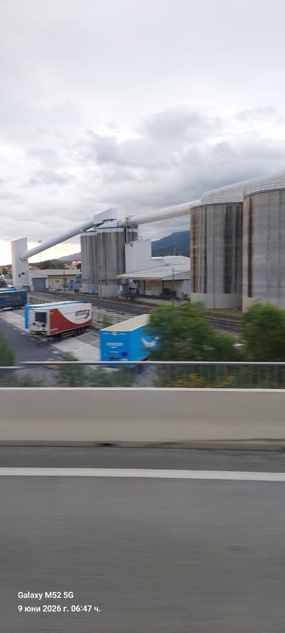 The image shows an industrial facility with large cylindrical silos and a long conveyor belt system. In the foreground, there are several trucks, including a red and white truck with "Saegens" written on it, and a blue truck with a white curved logo, possibly representing Amazon. The scene is captured from a moving vehicle, indicated by the motion blur of the foreground and the overall slight blurriness. The sky is overcast with clouds. The location is identified as Le Boulou, France. The bottom left corner of the image displays a timestamp: "Galaxy M52 5G 9 юни 2026 г. 06:47 ч.".