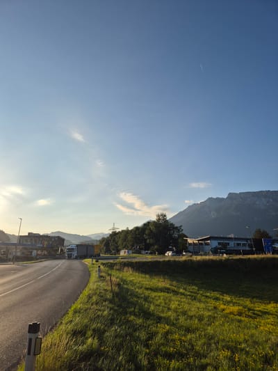 The image depicts a roadside scene in Niederndorf, Austria, bathed in the warm light of a setting or rising sun. A paved road curves from the lower left towards the center, bordered by a lush green grassy verge dotted with small yellow flowers. In the foreground, two reflective bollards stand sentinel along the road.
On the left side of the road, a modern building with prominent signage, likely a gas station or service area, is visible. A white semi-trailer truck is parked or driving on the approach to this area. Further in the background, trees and distant mountains form a scenic backdrop. A tall industrial tower with electrical wires stretches towards the sky behind the buildings.
On the right side of the road, a low-rise building with a modern facade stands. A blue traffic sign with international symbols is positioned near this building. The sky above is a clear, bright blue, with a faint contrail from an aircraft visible high up and wispy white clouds lower down. The overall impression is one of a serene, rural or semi-rural setting transitioning into evening or early morning. No people are clearly discernible in the image.