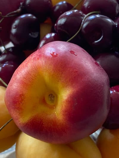 A close-up, overhead shot features a nectarine in the foreground, its skin a gradient of red and yellow, speckled with water droplets. It rests against a backdrop of dark, plump cherries, their surfaces shiny and reflective. The cherries are densely packed, with some stems visible. The overall impression is a still life of fresh fruit, highlighting the textures and colors of the produce. The setting, Askarovo, Russia, is noted but not visually depicted.
