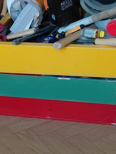 The image captures a detailed view of a cluttered indoor space, likely a storage area or workshop, with a distinctive background. The left side of the frame is dominated by an assortment of items, including various tools and components. A red bottle cap is visible near the top, alongside a clear plastic object with a yellow cap, possibly a small bottle or glue stick. Several wooden handles of tools are interspersed throughout the clutter. A prominent black object features white text clearly reading "18 VOLT" along with several warning icons depicted as crossed-out squares, suggesting it is a battery pack or part of a cordless tool. Other items include a clear cylindrical container filled with small wooden sticks, a stack of thin white rectangular pieces, parts of an orange tool, and red-handled scissors.
To the right, three distinct, brightly painted vertical boards create a colorful background or partition. From left to right, these boards are yellow, green, and red. They appear to be made of wood or a similar material, showing some wear and slight gaps between them. The very bottom right of the image reveals a wooden floor laid in a herringbone pattern, indicating an indoor setting. No people are visible, and no specific activity is taking place beyond the items being stored. The lighting suggests an indoor environment, but the time of day is not discernible. A faint, small "R" or similar mark is visible on the red board near the top.