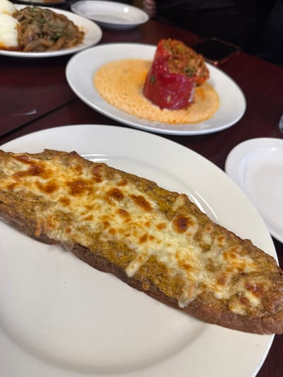 The image shows a table setting with three white plates on a dark wooden surface. On the right is a white plate with a baguette topped with melted cheese. The bread has a golden-brown crust and the cheese is bubbly and browned in places. In the center is another white plate with a stuffed red bell pepper atop a yellow puree. To the left is a smaller white plate with a portion of mushrooms and what appears to be mashed potatoes. A small white dish is also visible to the left. A portion of a person's dark clothing is visible in the upper left corner. Based on the presence of the stuffed pepper, the location may be the Allegretti's Bakery Cafe in Des Plaines, Illinois.