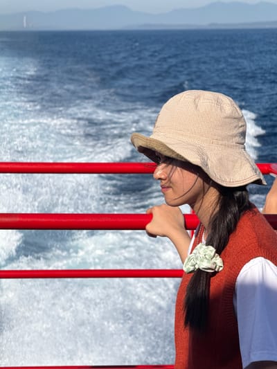 A young person with long, dark hair, wearing a bucket hat, an orange vest, and a white shirt, is leaning on a red railing while on a boat at sea. The person is looking out at the water, with their profile visible. The sea is blue and choppy, with white foam from the boat's wake. In the distance, mountains are visible on the horizon. The scene suggests travel or leisure on the water. The specific location in the city of Dong Son, Vietnam, cannot be determined from the provided image.