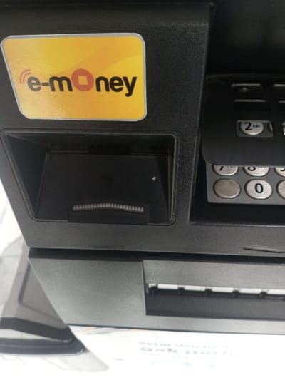 The image shows a close-up of an ATM. It features a card reader slot below a yellow "e-money" sticker and a numeric keypad to the right. The ATM is dark gray, and the keypad includes buttons for "2 ABC", "7 PRS", and "0". Part of another ATM's side and promotional material are visible in the lower portion of the frame.