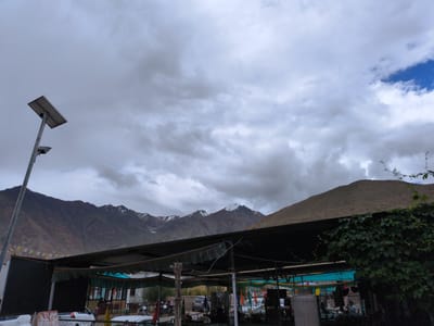 The image depicts a view of Khardung, India, under a cloudy sky. In the foreground, a black awning supported by poles covers a seating area with tables and chairs. Prayer flags are strung along the top edge of a dark structure to the left of the awning. Behind the awning, several vehicles are visible, including cars and motorcycles. Further in the background, a vast, barren mountain range dominates the landscape, with some snow-capped peaks visible. A solar-powered street lamp stands to the left of the frame, casting a shadow on the ground. The overall scene suggests a roadside establishment or rest stop in a mountainous region.