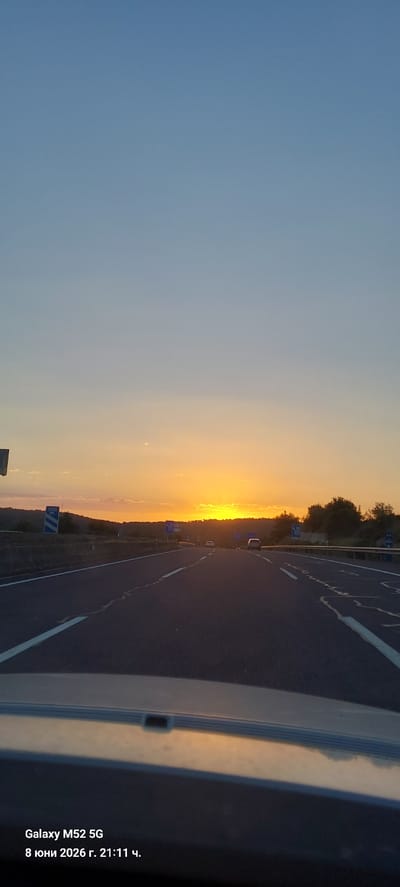 The image depicts a sunset over a highway, captured from the perspective of a vehicle. The sky transitions from a clear blue at the top to warm orange and yellow hues near the horizon, indicating the sun is setting. The highway stretches into the distance, with lanes marked by white lines. Several cars are visible on the road, traveling in the same direction. On the left side of the highway, there is a barrier and some vegetation, along with road signs. The foreground shows the dashboard and windshield of the vehicle. The bottom left corner of the image contains text indicating the device used to take the photo ("Galaxy M52 5G") and the date and time ("8 юни 2026 г. 21:11 ч."). While the image itself does not contain explicit geographical markers for the city of Canals, Spain, the timestamp format with Cyrillic characters suggests it may have been taken in a location where such a format is used, or the user has set their device to that language. Based on general knowledge of highway sunsets, this could be a scenic route in the region.