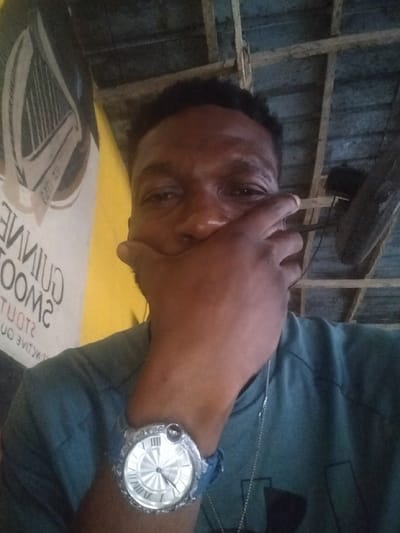A man covers his mouth with his hand, wearing a large wristwatch with Roman numerals and a silver chain necklace. A Guinness advertisement hangs on a yellow wall to the left. Above, a wooden slat ceiling is visible with an old fan attached. The man wears a teal shirt.