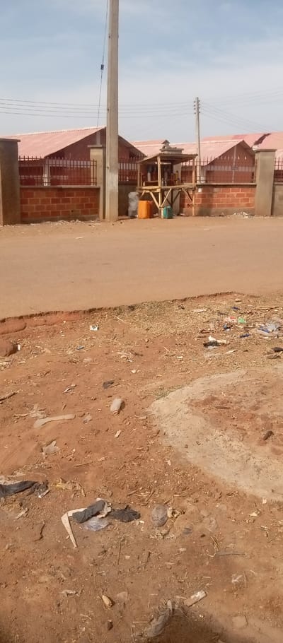 The image displays a daytime street scene in Kasuwan Magani, Nigeria. The foreground consists of a dirt and gravel ground surface extensively littered with various debris, including plastic bottles, bags, paper, and organic matter. This unpaved area borders a slightly elevated, reddish-brown paved road that traverses horizontally across the midground.
In the background, positioned beyond the paved road, a low brick wall topped with a black metal fence delineates a property. Behind this boundary, multiple single-story buildings with red corrugated metal roofs are visible under a clear, light blue sky.
A prominent utility pole, light gray in color, stands vertically on the roadside to the left of the image's center, extending out of frame at the top. Wires are attached to this pole. To its right, near the brick wall, is a small, elevated wooden kiosk with a simple canopy roof. Beneath the kiosk, an orange jerrycan, a green jerrycan, and a grey plastic bag are visible. The top of the kiosk holds numerous smaller containers, consistent with a roadside vending operation. Further to the right in the background, another utility pole with multiple insulators and power lines is present. No individuals or vehicles are depicted in the scene.