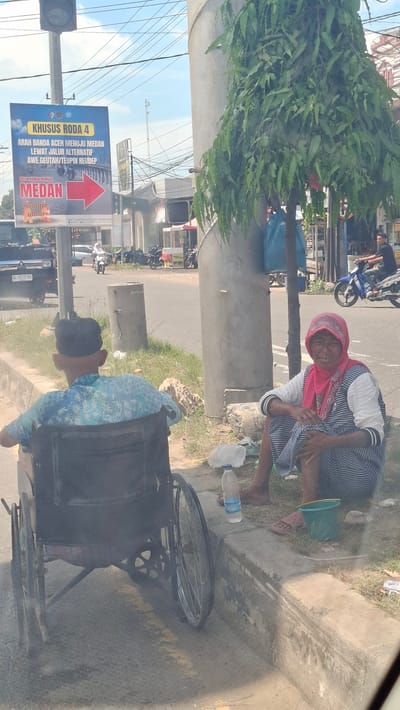 In Pantegajah, Indonesia, a man in a wheelchair faces away from the camera, his back to the street. He wears a blue patterned shirt and a black skullcap. To his right, a woman sits on the curb, facing the viewer. She is wearing a pink headscarf and a striped dress, and her legs are crossed. In front of her is a green bucket. A bottle of water and some trash are also on the ground near the curb. A large tree with green leaves shades the area. In the background, traffic can be seen on the street, including cars and motorcycles. A sign directing traffic to Medan is visible on the left side of the image. The scene appears to be a street corner with a sidewalk and a grassy area.