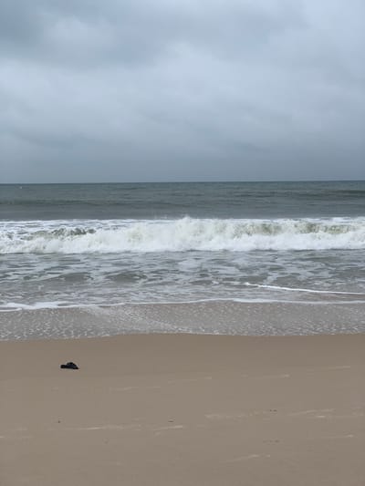 The image displays a wide view of a beach in Ibeju, Nigeria, under a predominantly overcast sky. The scene is bisected horizontally by the horizon line, separating the grey-blue ocean from the light grey, cloudy sky. Multiple waves are observed, with one significant white-crested wave breaking in the mid-ground, generating foam that extends towards the sandy shore. The foreground consists of light tan sand, exhibiting wet darker patches near the water's edge. A single, dark, amorphous object, possibly a shoe or discarded item, rests on the dry sand in the lower-left portion of the frame. No individuals, animals, or active human interactions are present.