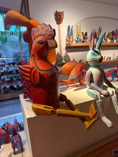 The image showcases a collection of vibrant, handcrafted alebrijes. A red-toned animal with intricate patterns dominates the foreground, while a colorful rabbit figure floats above, adorned with polka dots. Another winged animal, orange, sits between them. These folk art sculptures are displayed on a shelf, with more alebrijes visible in the background, some mounted on the wall, creating a lively and artistic scene.