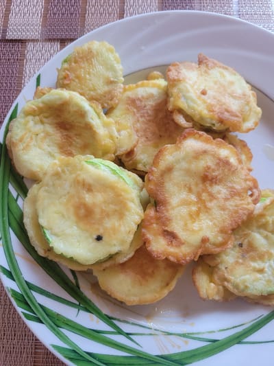 A plate of fried zucchini slices is presented on a table covered with a woven placemat. The zucchini slices are coated in a light batter and fried to a golden brown. Some slices have visible green edges, indicating they are zucchini. The plate itself is white with a green leaf design on the right side. The lighting suggests it is daytime. No people or text are visible in the image. The scene appears to be a home setting, possibly in Belaazyorsk, Belarus.