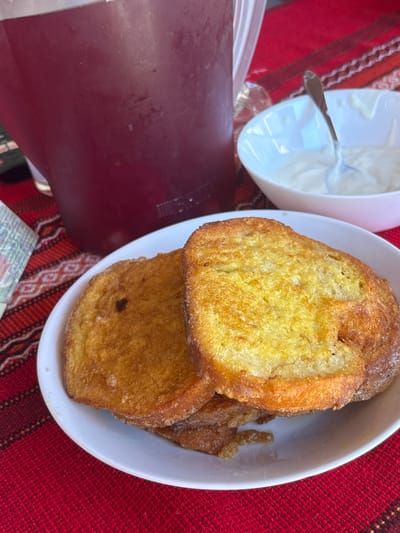 A close-up, high-angle shot shows a meal served on a red woven placemat with a geometric pattern. In the foreground, a white bowl contains two slices of golden-brown French toast, with a third slice partially visible beneath them. To the left of the bowl is a large, clear pitcher filled with a dark red beverage. In the background, a smaller white bowl holds a white, creamy substance, likely yogurt or a similar dairy product, with a spoon resting inside. The scene appears to be an indoor setting, possibly a home or restaurant in Enevo, Bulgaria, given the placemat's traditional textile design often found in the region. There are no people visible in the image.