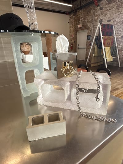The image displays an indoor scene, likely a retail or gallery space, featuring an eclectic collection of objects arranged on a metallic counter or display surface. The foreground is dominated by several striking items.
On the metallic surface, there are two large, transparent, and textured blocks resembling concrete cinder blocks, but made of a clear, possibly frosted glass or acrylic material. One block stands prominently in the center, and another similar, slightly smaller block is positioned to its left. A shiny silver chain is draped over the central transparent block, with one end attached to a silver metallic object, which appears to be a bag or container with a gold-colored clasp and rings. The chain features a small hook-like closure at its free end. To the far left, a black baseball cap with "BOND" embroidered in white is partially visible. To the right of the transparent blocks, a small, light-colored miniature concrete block, similar in shape to a cinder block, rests on the metallic surface. Text is visible on its side, reading "CAOS MORTAR" and "BOND".
The background reveals an exposed brick wall, characteristic of an industrial or repurposed building. Industrial pipes run along the wall. A white door with a silver handle is set into the brick, and above it, a red "EXIT" sign with white lettering is illuminated. A black sign with unreadable white text is also mounted on the wall above the door. Further in the background, a white metal shelving unit holds various folded textiles, possibly rugs or blankets, displaying different patterns and colors (red and black geometric, yellow, and green patterned). The overall setting suggests a modern, urban aesthetic in what appears to be a store or showroom in the City of Hudson, United States of America. No people are visible, and the scene is lit by artificial indoor lighting.