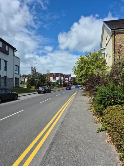 The image captures a street scene in Central Park, Ireland, on a day with a partly cloudy sky. A two-lane asphalt road runs through the frame, bordered by a sidewalk. Yellow double lines are painted on the road, indicating no passing. Several cars are visible on the road, including a dark sedan parked on the left and a black sedan further down on the right, along with two blue cars driving away from the camera.
Buildings line both sides of the street. On the left, a modern white building with grey stone accents is prominent. On the right, a building with a cream-colored facade and brick accents is visible, with lush green and reddish-purple foliage in front of it. In the distance, a church spire with a clock is visible above the rooftops, along with more traditional-looking buildings, one of which features a distinctive red and white design. Orange traffic cones are placed along the sidewalk on the right side of the street. The overall impression is a residential or mixed-use area with a blend of contemporary and older architecture.