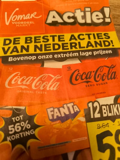 The image is a close-up of a flyer from a Dutch supermarket. The flyer advertises "De Beste Acties van Nederland" (The Best Deals in the Netherlands) with the slogan "Bovenop onze extreéém lage prijzen" (On top of our extremely low prices). It prominently features promotions for Coca-Cola (both original taste and zero sugar), Fanta, and a product labeled "12 BLIKH" which likely refers to a 12-pack of cans. There is also a section indicating "TOT 56% KORTING" (UP TO 56% DISCOUNT). The flyer has a vibrant orange and yellow color scheme. The supermarket's name, "VOMAR VOORDEEL MARKT," is visible in the top left corner. The flyer appears to be dated "zondag 28 juni t/m zaterdag 4 juli 2026," indicating the sale period. The image is cropped in a way that suggests it's part of a larger advertisement or leaflet, possibly found in a newspaper or directly mailed to households. The specific location within Almere, Netherlands is not determinable from the image content alone.