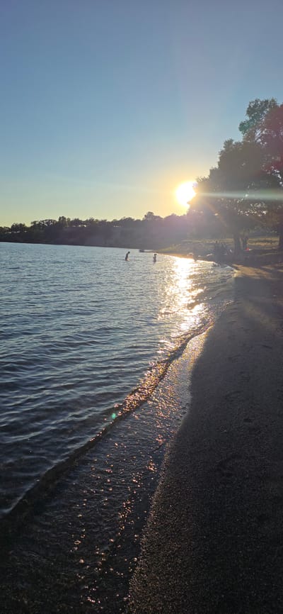 The image depicts a serene sunset scene at a beach on the shore of Issyk-Kul Lake in Kyrgyzstan. The sun, low in the sky, casts a golden glow across the water, creating a shimmering path of light. Gentle waves lap at the sandy shore, where a few figures are visible. Two individuals are wading in the shallow water, appearing to be children. Further down the beach, a group of people are gathered, some seated and others standing near a large tree. The background shows a line of trees and hills under a clear blue sky. The overall atmosphere is tranquil and peaceful, suggesting a warm summer evening.