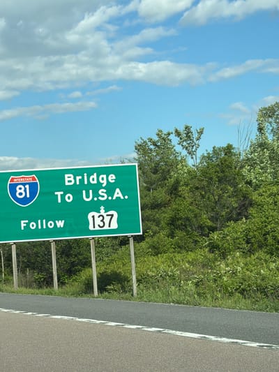 A green highway sign reads "INTERSTATE 81 Bridge To U.S.A. Follow 137". The sign is supported by two wooden posts and is set against a backdrop of trees and a blue sky with clouds. The sign is positioned next to a road.