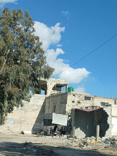 An outdoor scene in Al-Qadam Municipality, Syria, depicts a damaged urban environment under a clear blue sky with scattered white cumulus clouds. A large deciduous tree with dense green foliage occupies the left foreground, partially obscuring a light-colored masonry building.

In the midground, a multi-story building constructed of light-colored cinder blocks exhibits extensive structural damage. The upper floor and sections of the lower floor are missing walls, exposing interiors, and windows are either absent or broken. A green cylindrical water tank is visible on the roofline. Adjacent to this structure, a smaller building, also damaged, features a reddish-brown sheet metal roof section and exposed masonry. Both structures show multiple impact marks consistent with bullet holes or shrapnel.

At ground level, directly in front of the damaged buildings, an unpaved area of dirt and debris leads to a makeshift shelter constructed with a white tarp roof supported by poles. Within this shelter, two adult males are observed. One individual, dressed in dark attire, is bent over a white motorcycle with a red seat, suggesting an activity involving its maintenance or repair. A second red motorcycle is also present, and another person, similarly dressed in dark clothing, is seated nearby. Miscellaneous objects, including several tires, are visible within and around the shelter.

The foreground consists of an unpaved path with scattered rubble, plastic bags, and general refuse. A thin, dark utility line traverses the upper right portion of the sky.