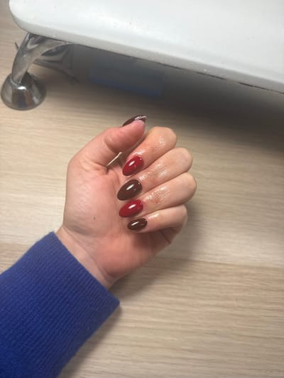 A close-up photograph depicts a human left hand, oriented with fingers pointing towards the upper right, resting on a light-toned wooden surface within an indoor establishment in Aix-en-Provence, France. The hand's fingernails are manicured with an oval/almond shape. The index and ring fingers are coated in a glossy dark red polish, while the middle and pinky fingers feature a glossy dark brown polish. The thumb nail is partially visible, displaying a dark base with a distinct white tip. The skin around the cuticles and on the fingers exhibits a moist sheen, indicative of a recent cosmetic application. A small, faint blue dot is visible on the dorsum of the hand. The wrist is covered by a textured blue fabric sleeve, likely knitted. In the upper left background, a portion of a white, rectangular device with a rounded edge is partially visible, supported by a metallic, reflective stand. The surface of this device appears to have a layer of particulate accumulation. The wooden surface beneath the hand displays a consistent linear grain pattern.