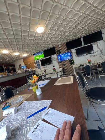 The image depicts the interior of a betting establishment in Bulgaria. The perspective is slightly askew, showing a table with betting slips, a pen, an ashtray, and a container with "Inbet" branding. A person's hand is visible, holding some of the betting slips. The background features a wall with a decorative pattern, several televisions displaying sports events and betting odds, and chairs typical of a betting shop or café. The location is likely in the city of Касапите, based on the OCR results on betting slips.
